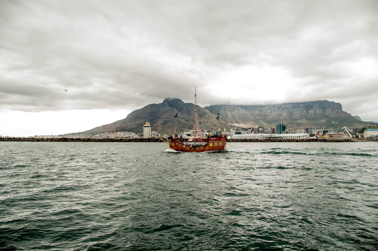 Wooden ship sailing in Cape Town with Table Mountain in the background under cloudy skies.